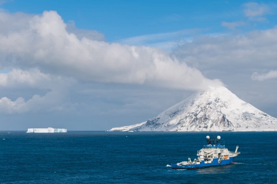 a research vessel sails in subantarctic waters near a snowy, steaming volcanic island