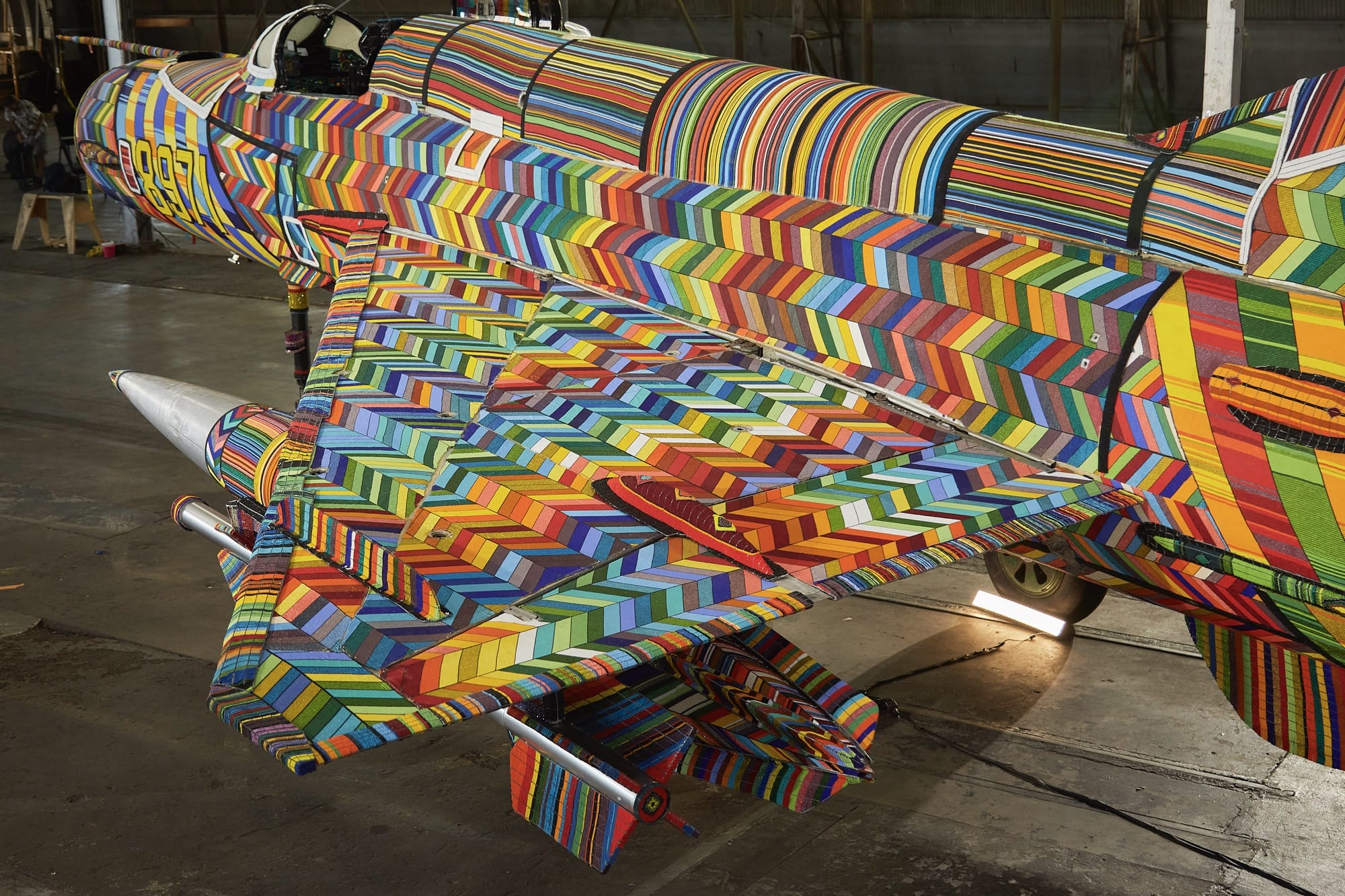 an overview of one wing and side of a fighter jet coated in colorful beaded patterns, sitting inside a hangar or warehouse